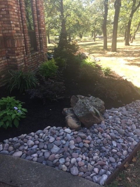 Brick building next to a garden bed with rocks, dark mulch, and plants, outside with trees.