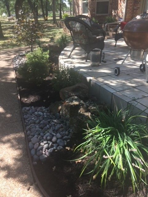 Stone-bordered garden bed with rocks, dark mulch, and various plants next to a patio with wicker furniture and a grill.