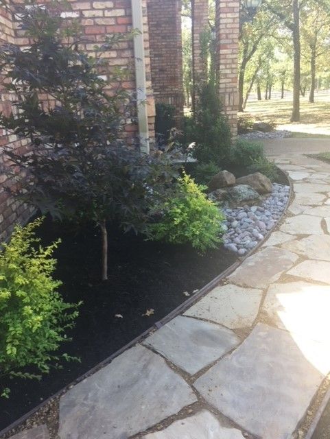 Landscaped garden bed with stone path, featuring various green shrubs and dark mulch. Brick wall in background.
