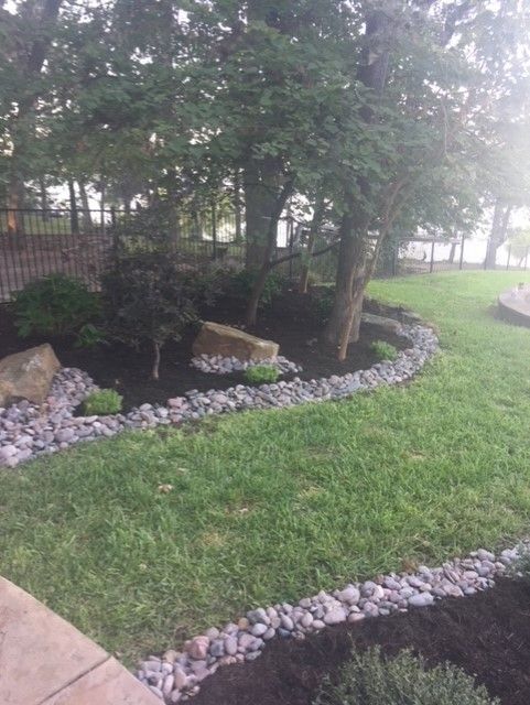 Lush green lawn with dark mulch beds bordered by gray river rocks, a small tree, and larger boulders.