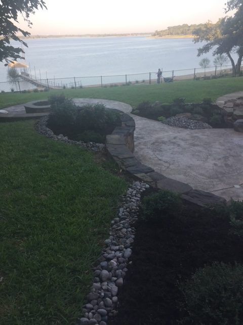Lush green lawn slopes toward a lake, with stone path and landscaping. A person stands by the water.