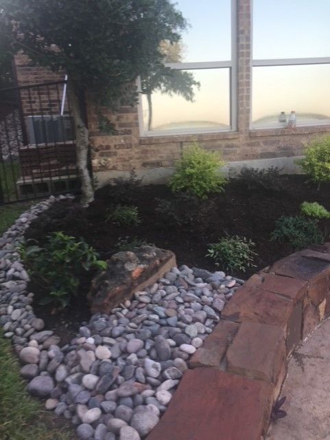 Landscaped garden bed with dark mulch, gray river rocks, and greenery in front of a brick wall.