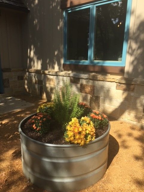 A metal stock tank planter with yellow, orange, and green flowers outside a building with a blue-trimmed window.