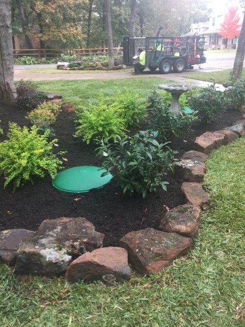 Garden bed with dark mulch, green plants, rock border, and a bird bath. Landscapers in the background.