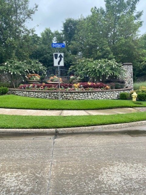 Entrance to a neighborhood with a brick wall, flowers, and a street sign. Cloudy sky.