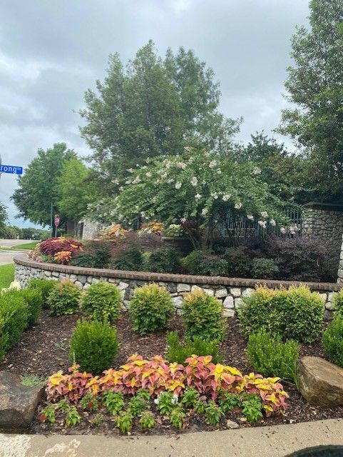Landscaped entrance with stone wall, flowering trees, shrubs, and colorful coleus. Cloudy sky.
