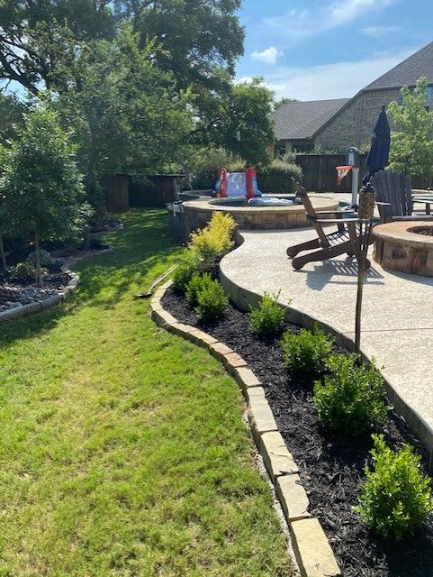 Lush backyard with stone border, plants, and a concrete patio area with a fire pit, chair, and play structure.