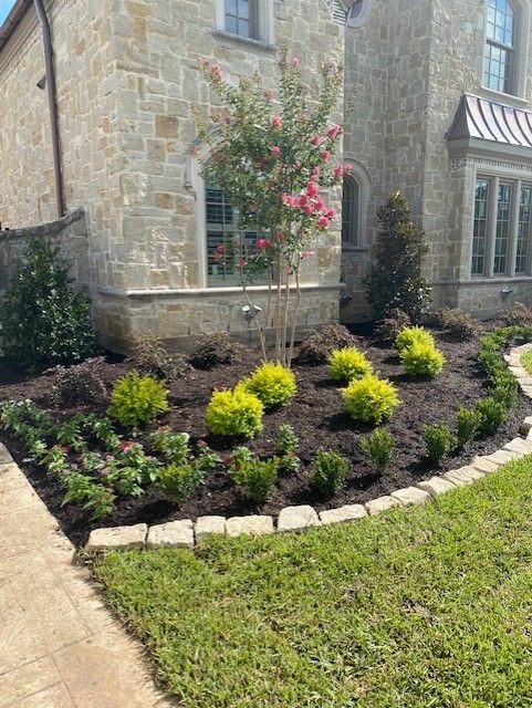 Stone house with a landscaped front yard featuring colorful flowers, shrubs, and a crape myrtle tree.