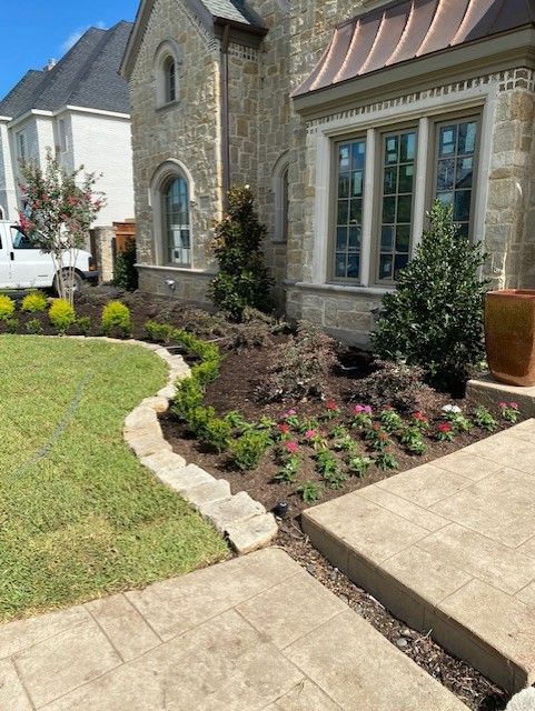 Stone house with landscaped flowerbeds, green grass, and a walkway.