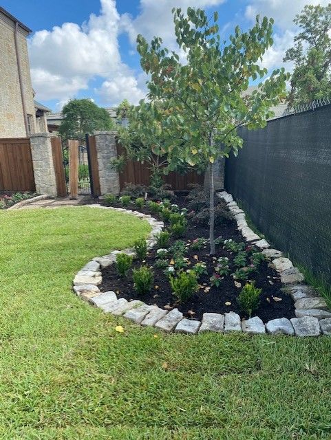 A landscaped garden with a tree, bushes, and flowers, bordered by stone and a green lawn.