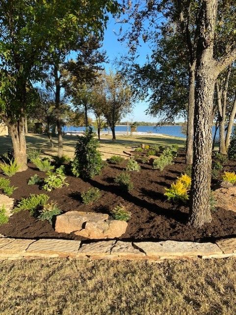 Garden bed with trees, lake in background; brown mulch, green plants, blue sky.