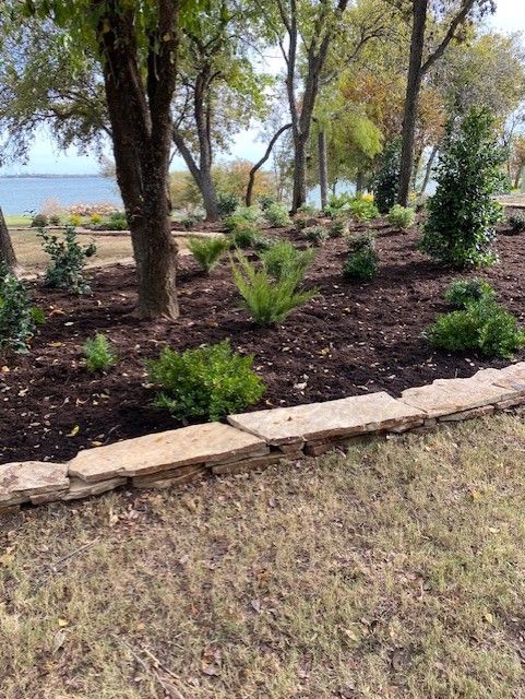 A landscaped garden bed with mulch and plants, bordered by stone, near a body of water.