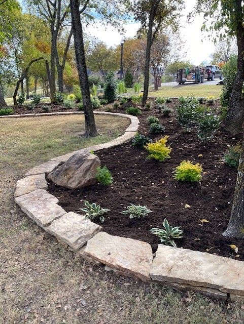 Stone-edged flower bed with plants and dark soil, in a park-like setting with trees and grass.