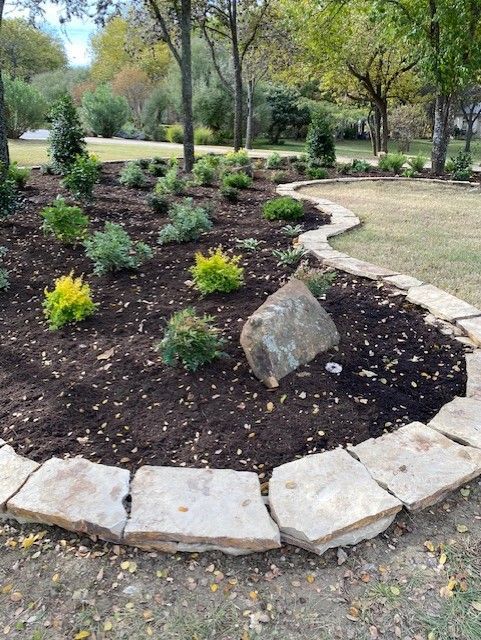 A landscaped garden bed with a stone border, filled with plants, mulch, and a large rock.