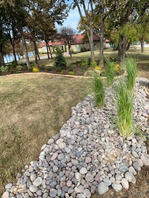A grassy area with river rocks and tall grass, with trees and a building in the background.