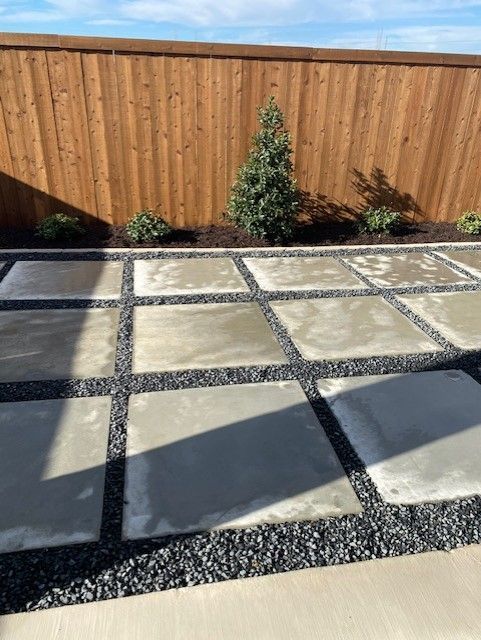 Concrete patio tiles with dark gravel in a backyard, bordered by a wooden fence and plants.