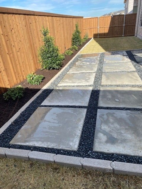 Concrete patio with black gravel accents, wooden fence, and small trees in backyard.