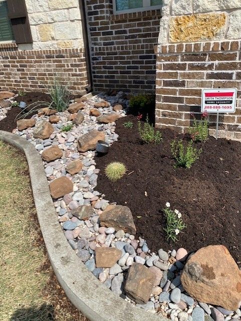 Landscaped garden bed with rocks, mulch, and plants in front of a brick house.
