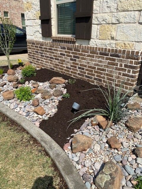 Front yard landscaping with rocks, mulch, and plants in front of a brick wall and window.