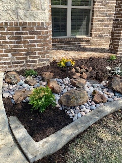 Landscaped flower bed with rocks, mulch, and plants near a brick building.