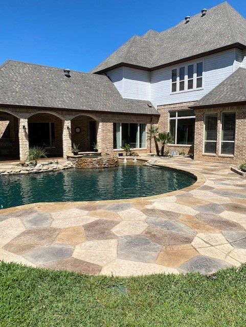 Backyard with a freeform pool, stone patio, and a two-story brick house under a clear blue sky.