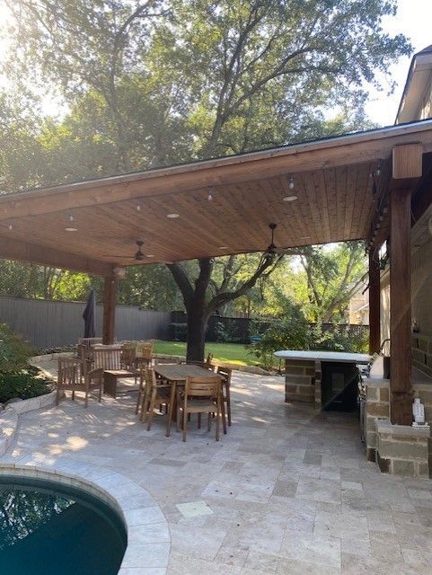 Outdoor patio with wooden beams, dining set, and built-in grill under a roof, near a pool.
