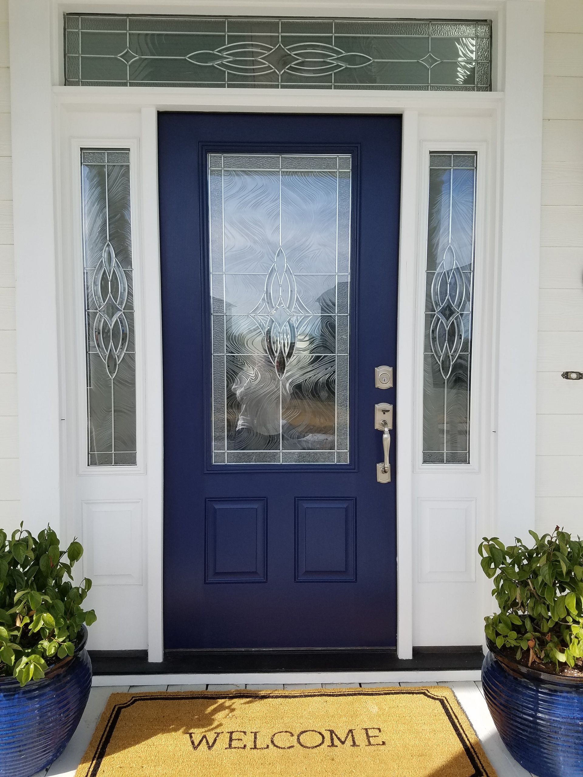 Blue front door with sidelights and transom, framed by white trim and potted plants on the entryway