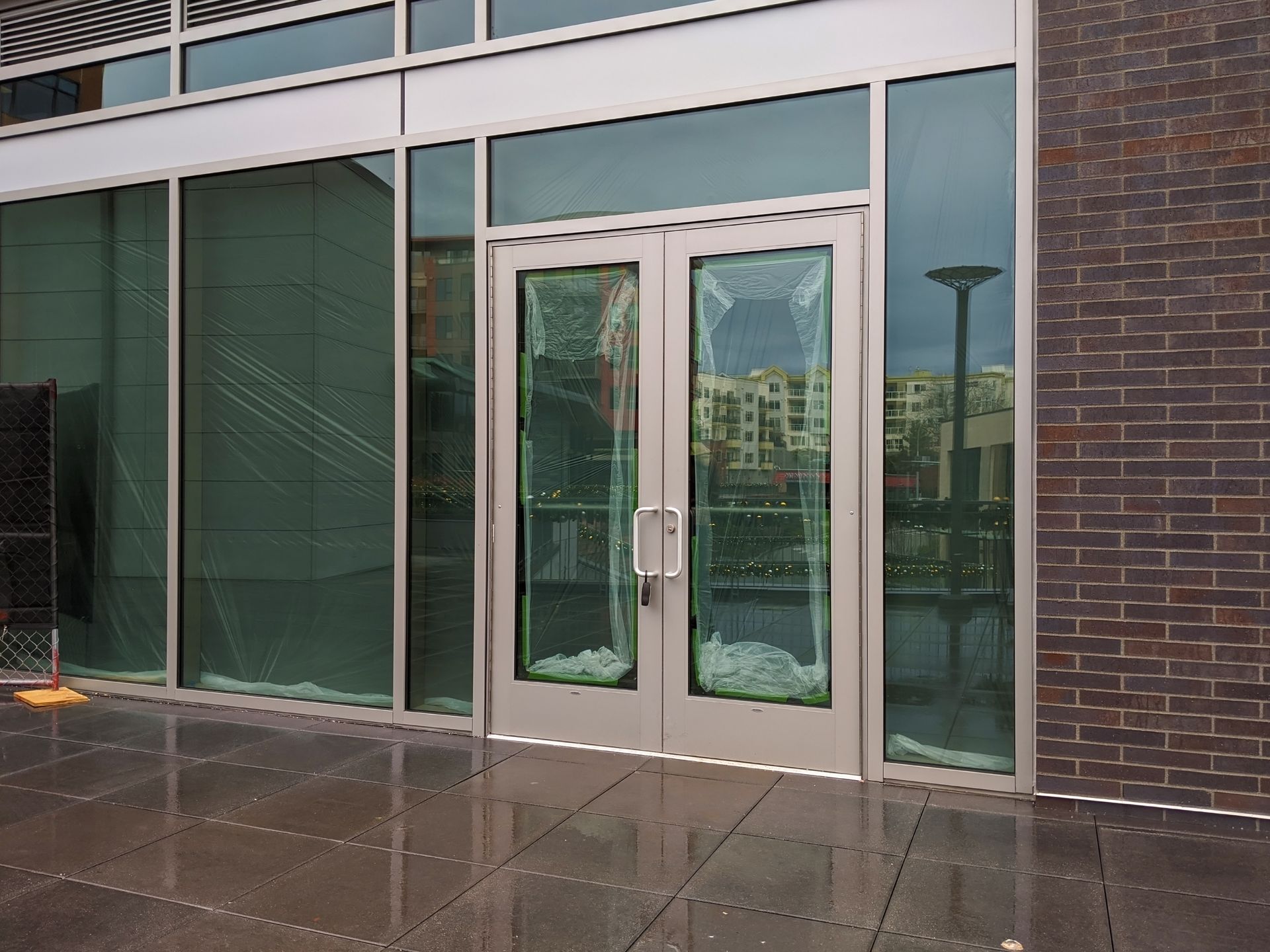 Glass double doors in a brick building entrance with wet pavement and green-tinted windows