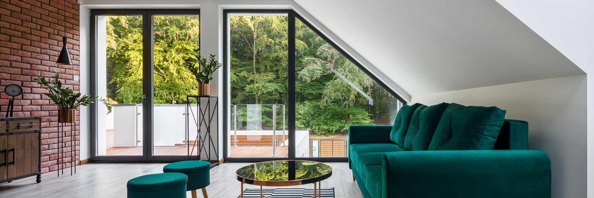 Bright loft living room with teal sofa, brick wall, skylight, and glass doors opening to a garden.