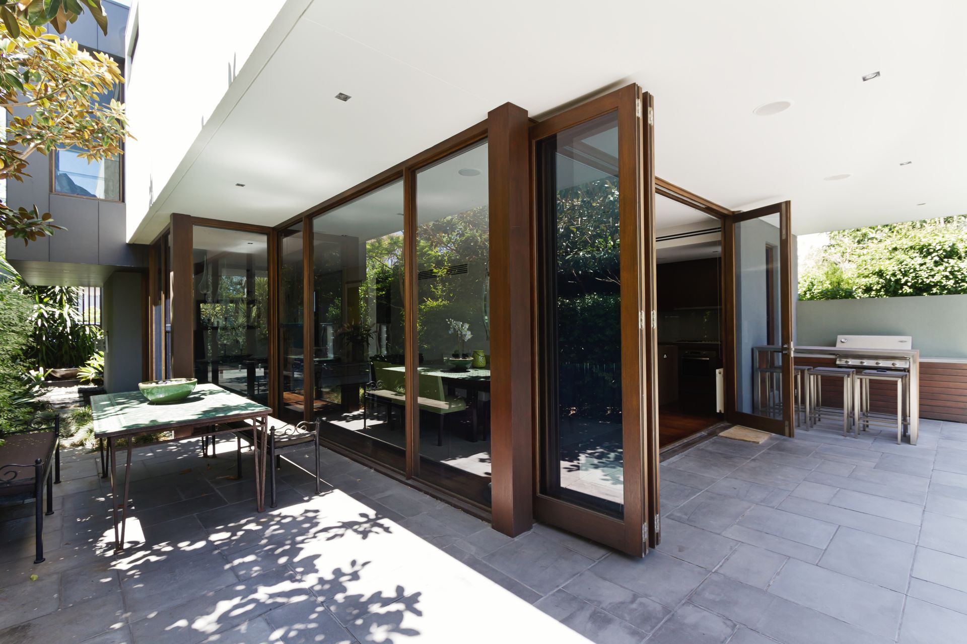 Modern patio with open glass doors, wooden frames, outdoor dining table, and sunlit stone flooring