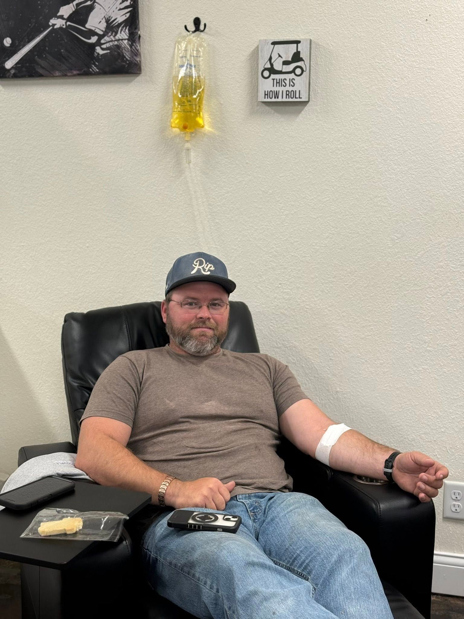 Man in chair receiving IV fluids, wearing hat and casual clothes, arm bandaged, indoors.