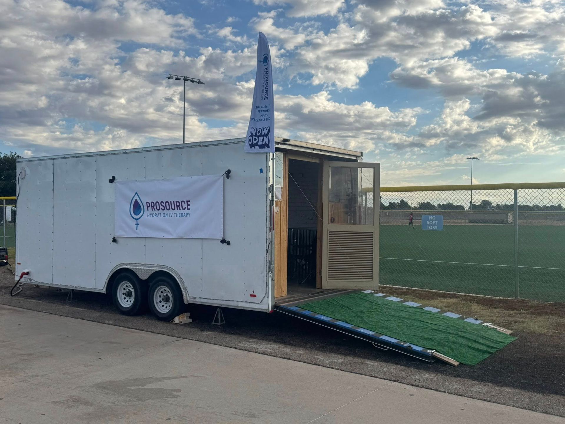 White trailer with a ramp open, next to a baseball field, under a cloudy sky.