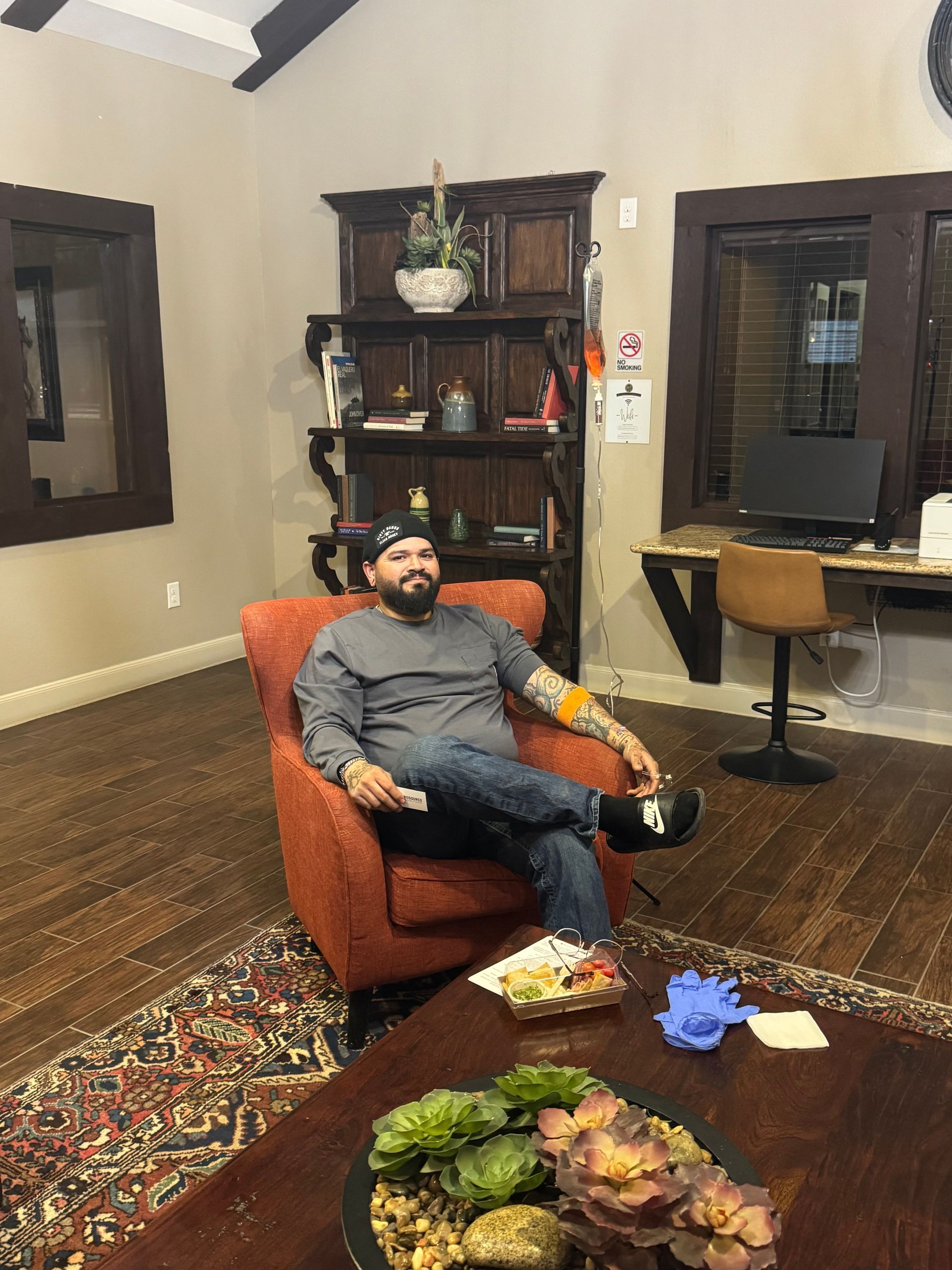 Man sitting in a red chair in a waiting room, with tattoos, holding something, looking relaxed.