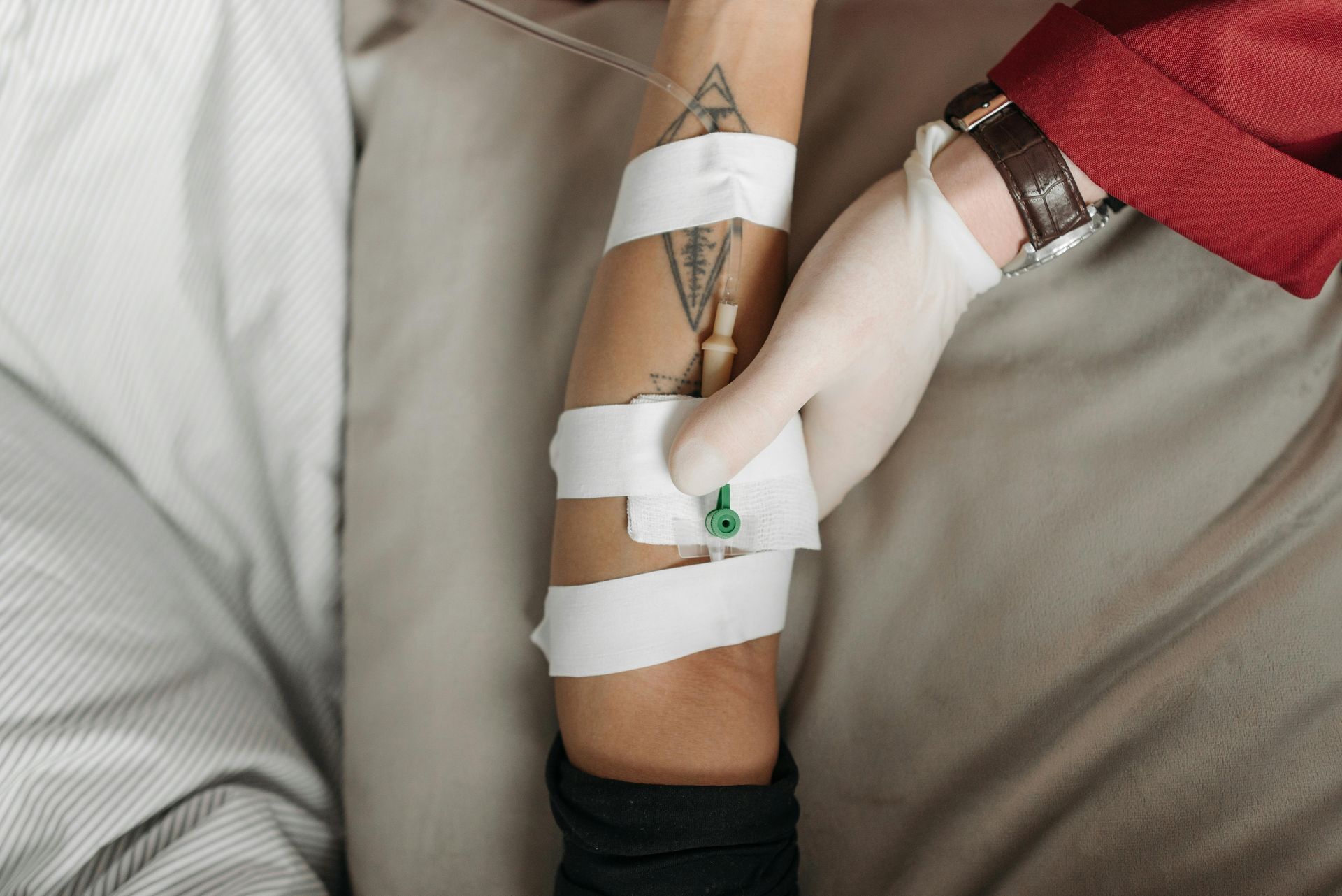 Person's arm with IV insertion, held by gloved hand. Bandages secure the needle.