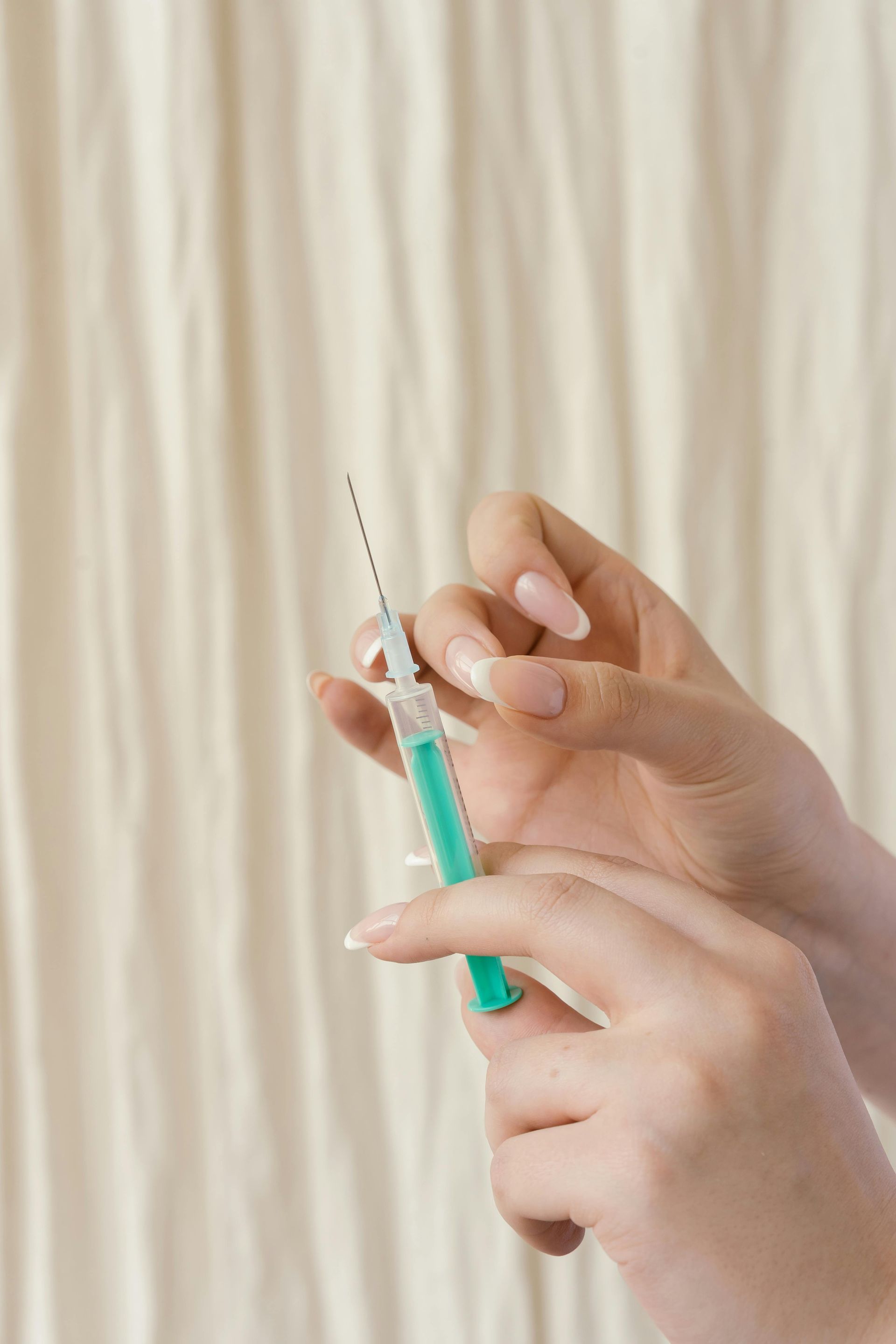 Hands holding a green and white syringe, preparing for an injection. Cream-colored background.