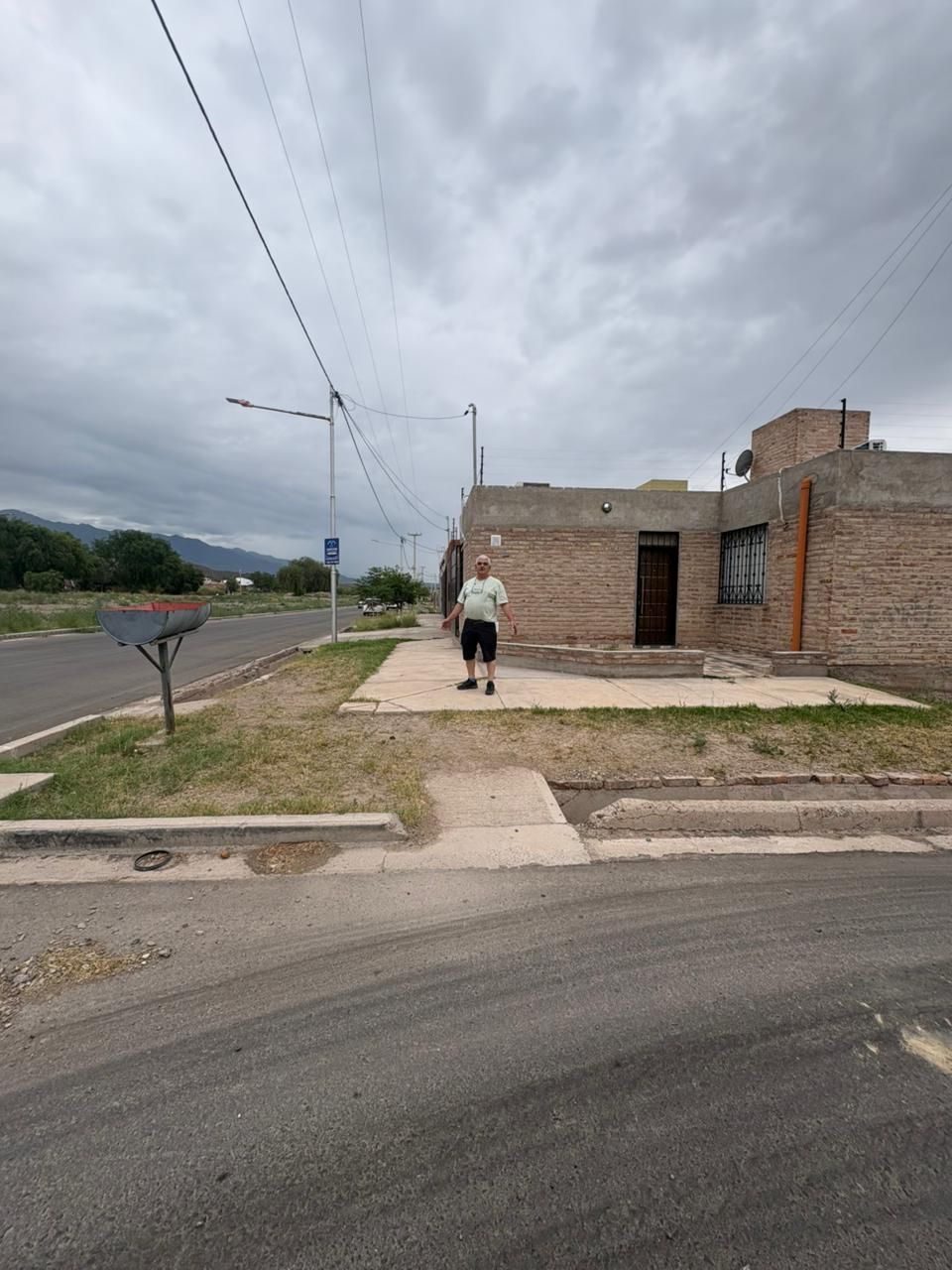 Hombre de pie frente a un edificio de ladrillos en un día nublado. Carretera vacía, líneas eléctricas y montañas visibles.