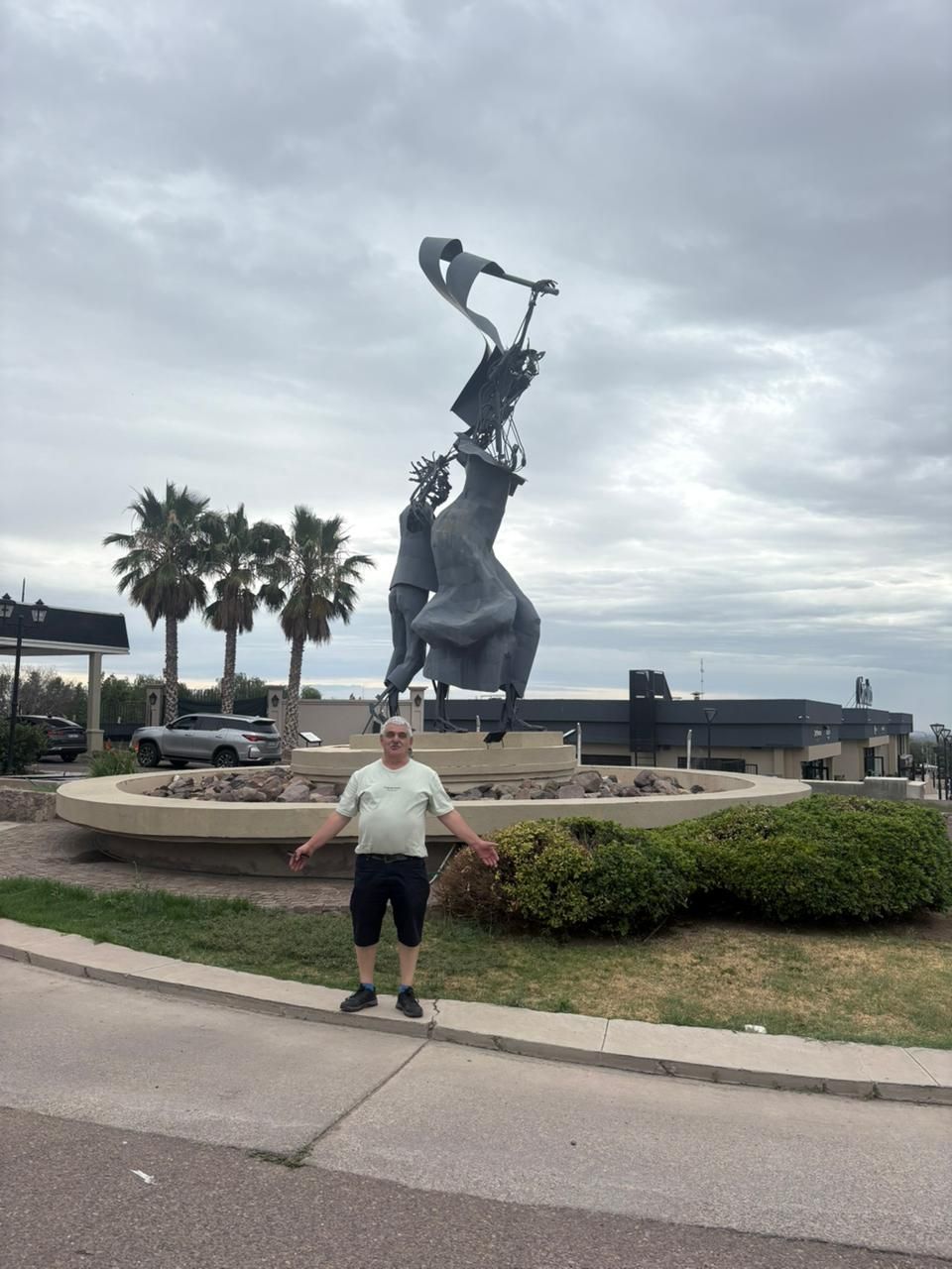 Hombre de pie frente a una escultura abstracta en una fuente. Cielo nublado y palmeras al fondo.