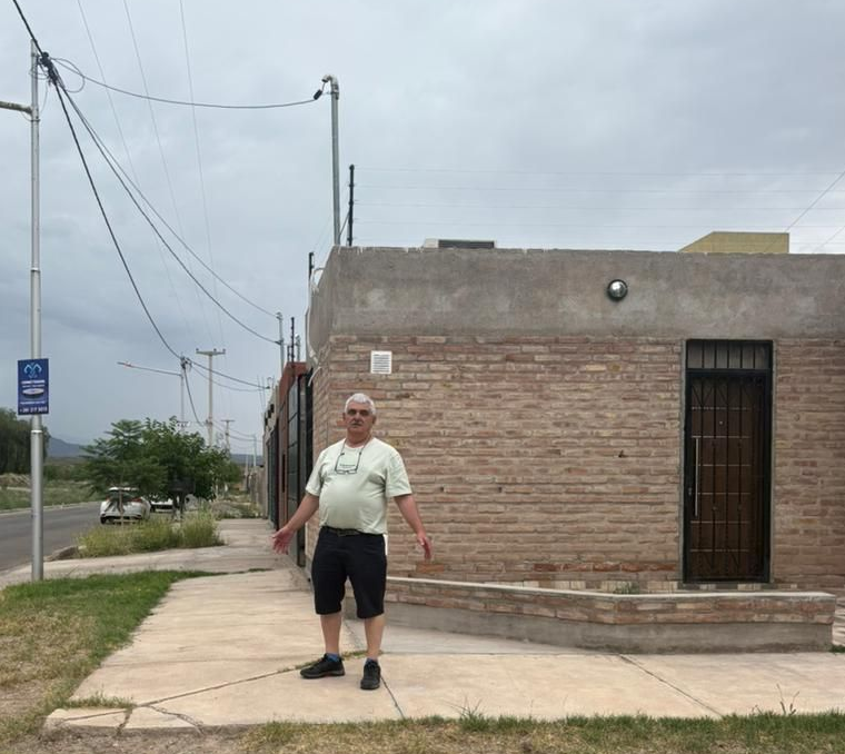 Hombre de pie en una acera frente a un edificio de ladrillos, con los brazos extendidos.