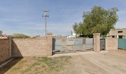 Muro de ladrillo con portón metálico gris. Camino de tierra. Poste telefónico. Árbol a la derecha.
