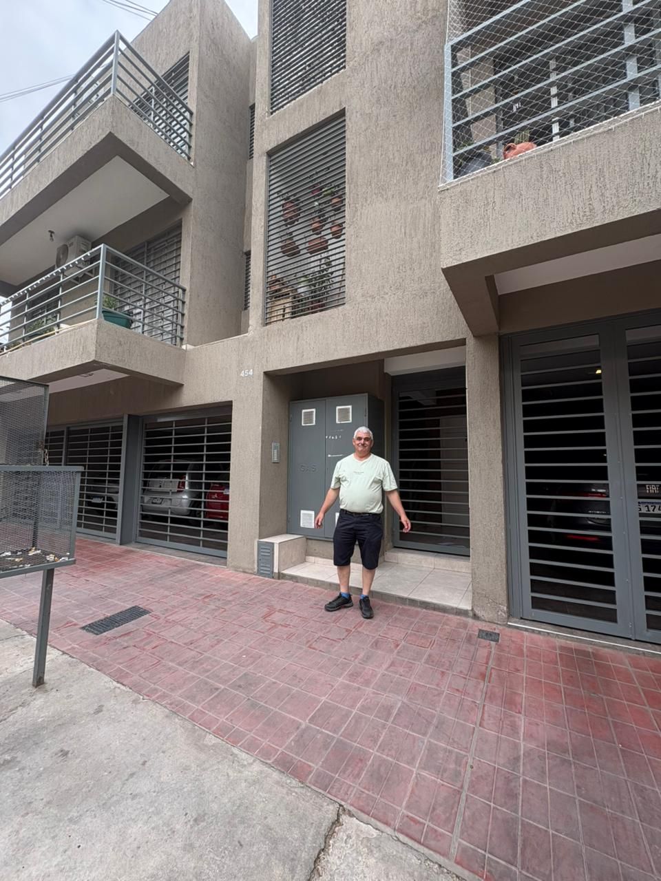 Hombre de pie frente a un edificio de apartamentos con puertas de seguridad. Pavimento de ladrillo rojo al frente.
