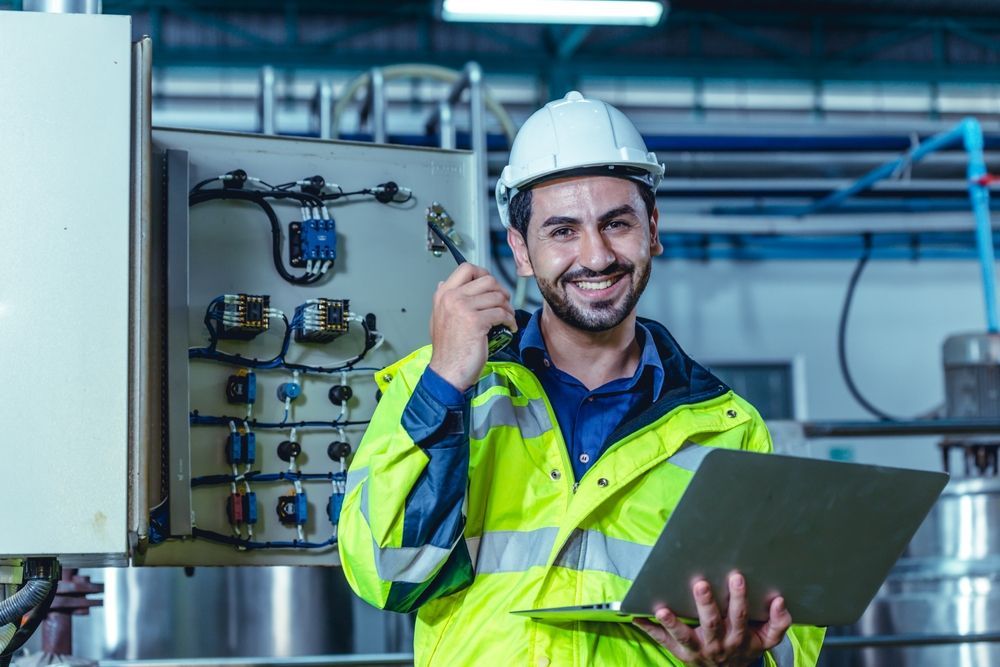 A man in a hard hat is holding a clipboard and a wrench in a factory.