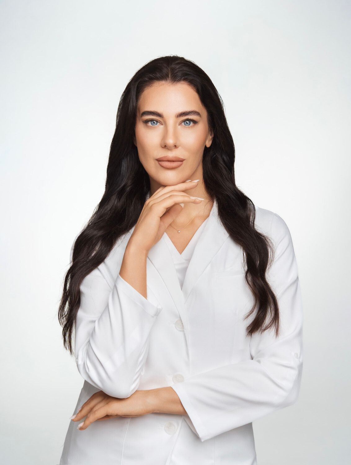 Frau mit langem dunklem Haar, trägt einen weißen Kittel, berührt mit der Hand ihr Kinn und blickt den Betrachter an.