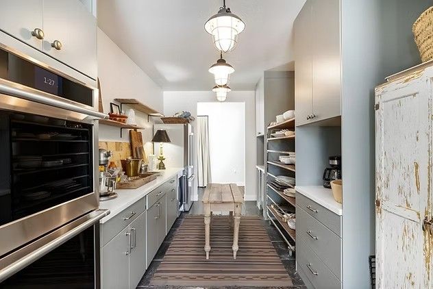 Narrow kitchen galley with white cabinets, stainless oven, open shelving, and a small table by the back door