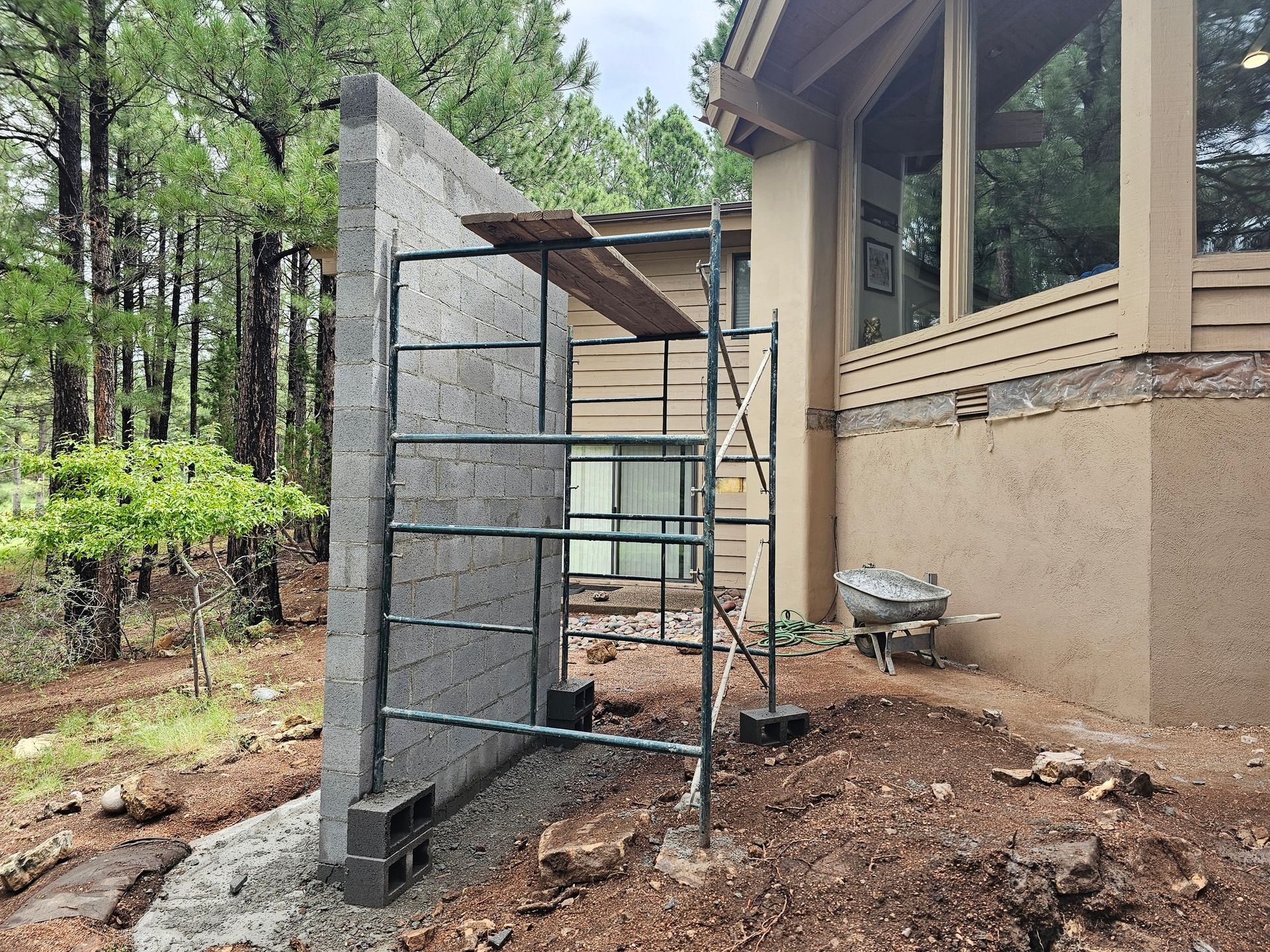 Scaffold beside a house under construction, with a tall gray retaining wall and wooded yard.
