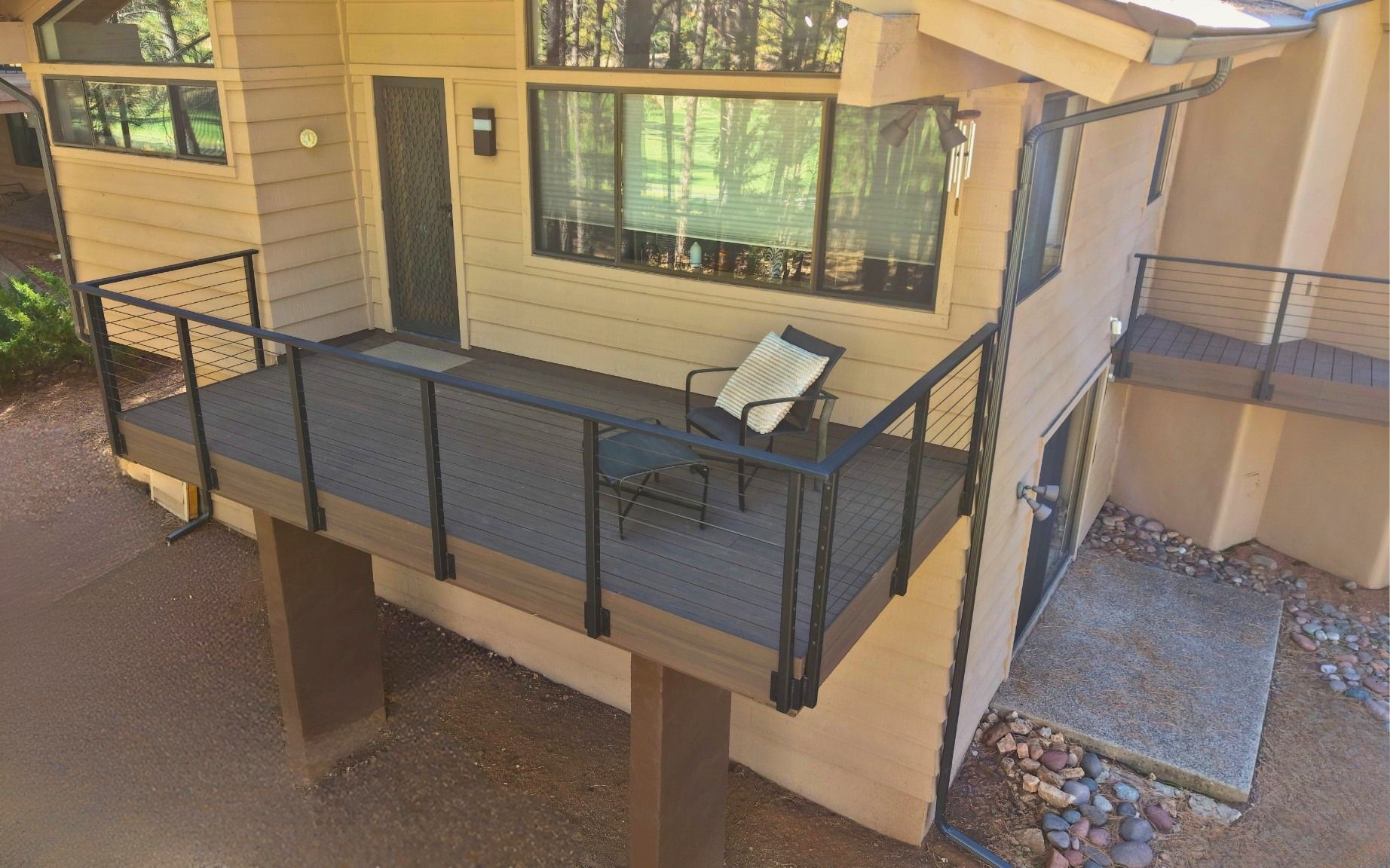 Balcony deck with black railings, two lounge chairs, and a large window beside a beige house