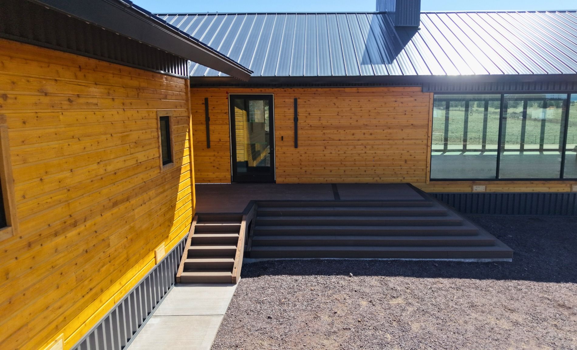 Modern wooden building entrance with steps, dark deck, and large windows under a metal roof