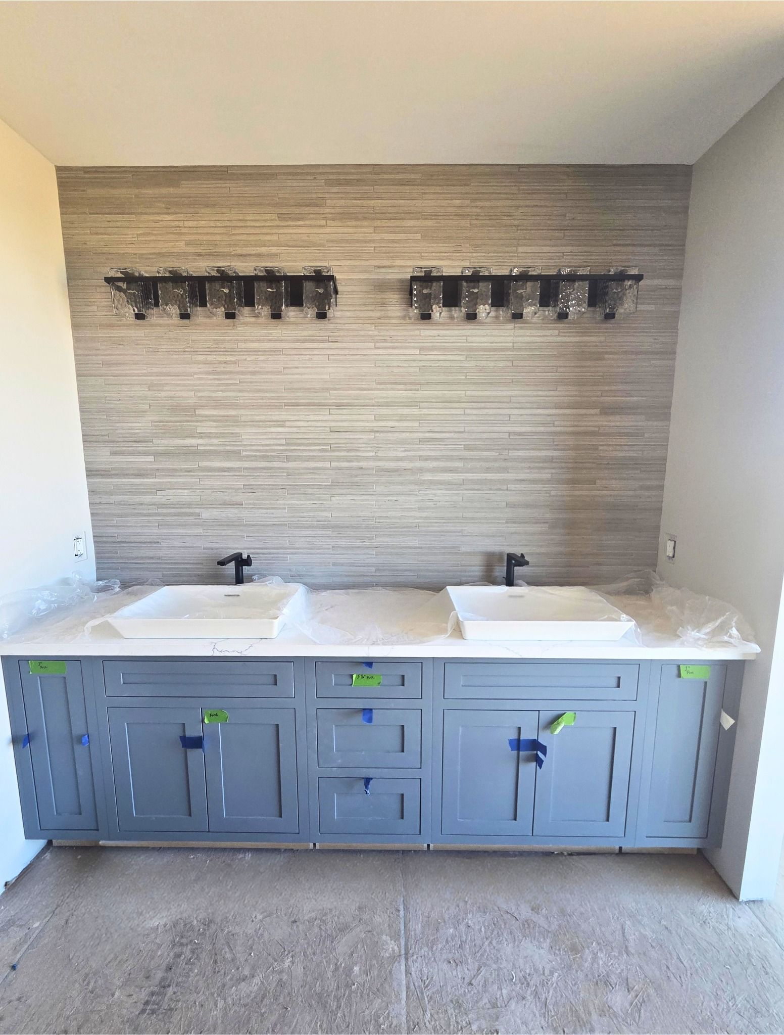 Modern bathroom vanity with two sinks, gray cabinets, and unfinished wall tile behind it