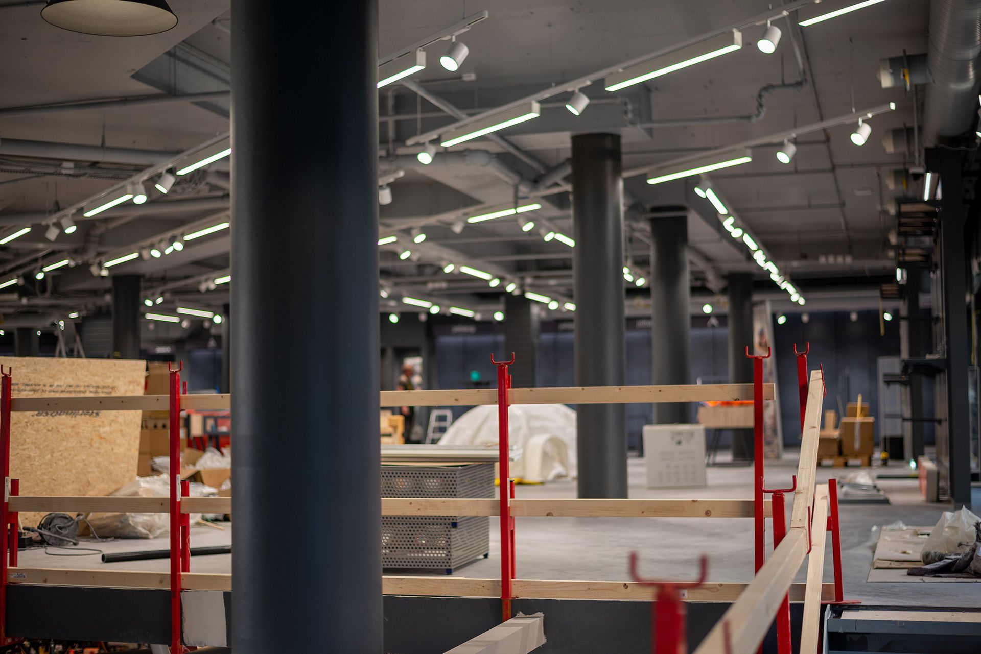 Indoor construction site with red safety posts, black columns, and overhead track lighting in a large open space