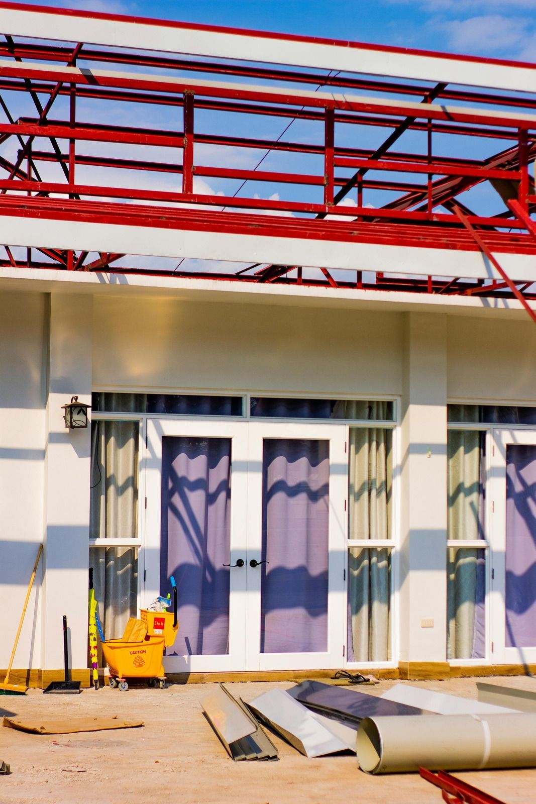 Construction site with red steel roof framing above glass doors and scattered building materials below