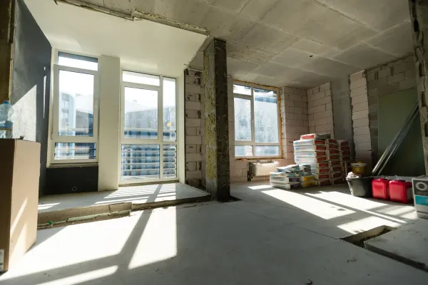 Unfinished apartment interior with large windows, exposed walls, and stacks of building materials in sunlight
